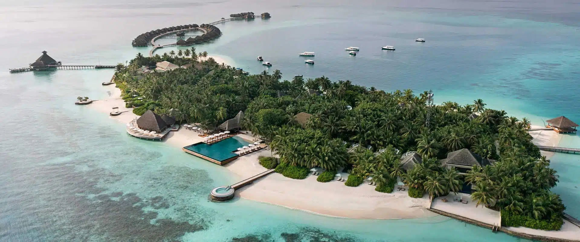 An aerial shot of Huvafen Fushi resort, Maldives. The island is surrounded by a clear blue lagoon and dotted with palm trees. Several small boats are anchored near the shore.